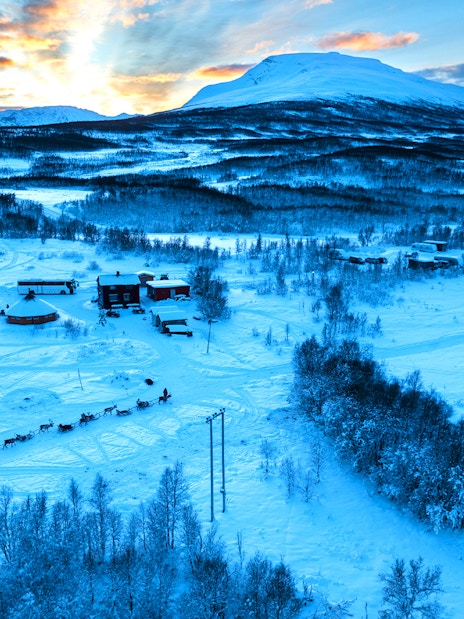 Reindeer sledding through snowy landscape with Sami tents and mountains in the background.