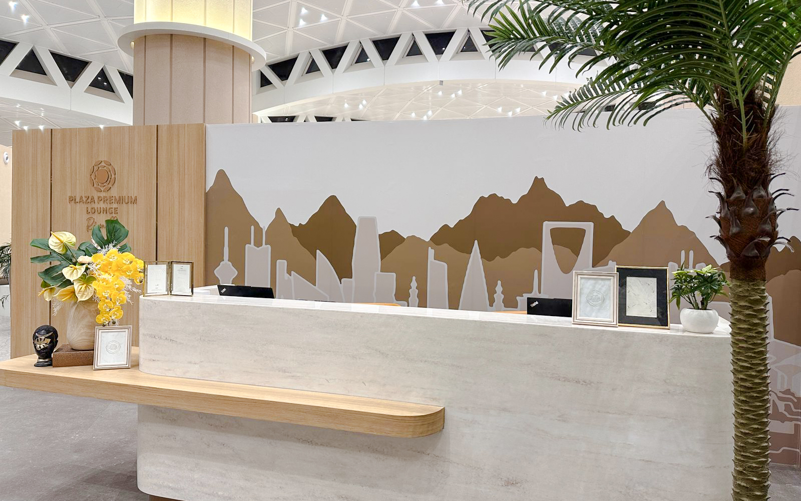 Reception desk at Plaza Premium Lounge, Riyadh, with decorative wall art and plants.