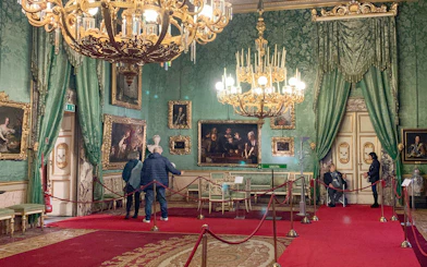 Palatine Gallery in Pitti Palace, Florence, with visitors viewing ornate paintings and chandeliers.