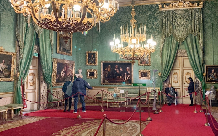 Palatine Gallery in Pitti Palace, Florence, with visitors viewing ornate paintings and chandeliers.