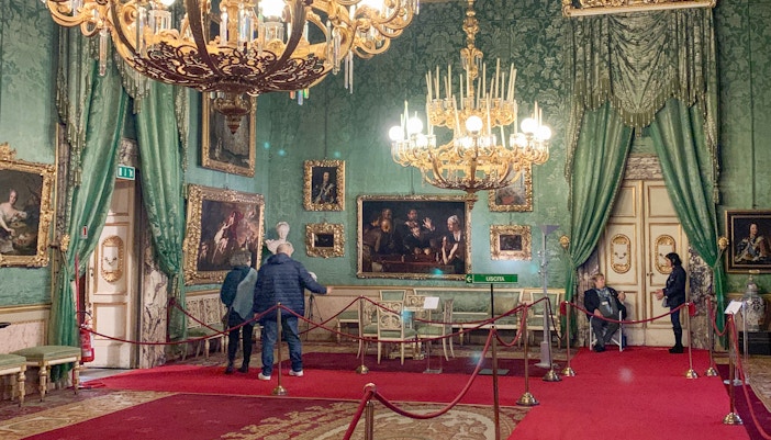 Palatine Gallery in Pitti Palace, Florence, with visitors viewing ornate paintings and chandeliers.