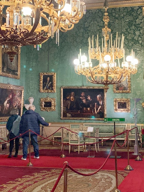 Palatine Gallery in Pitti Palace, Florence, with visitors viewing ornate paintings and chandeliers.