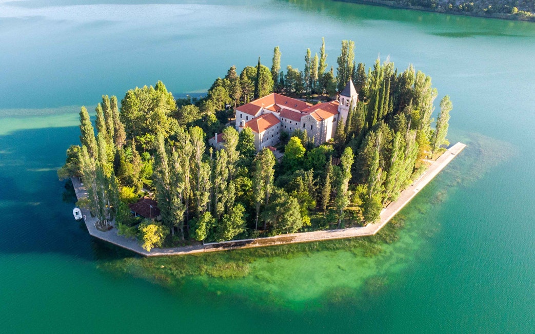 Aerial view of Krka Monastery surrounded by lush trees and turquoise water in Croatia.