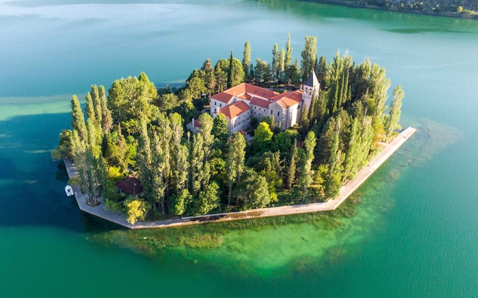 Aerial view of Krka Monastery surrounded by lush trees and turquoise water in Croatia.