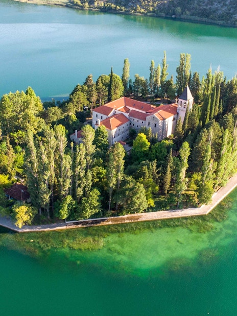 Aerial view of Krka Monastery surrounded by lush trees and turquoise water in Croatia.