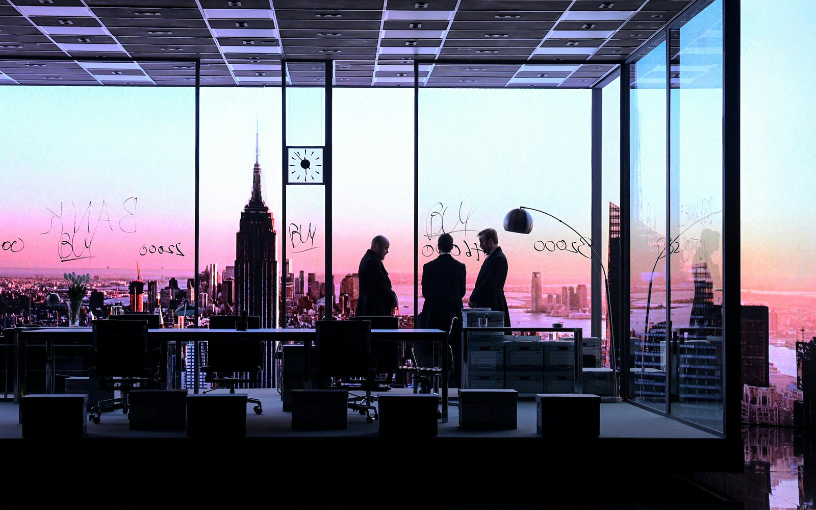 Three people in a modern office with a view of the New York City skyline at sunset, related to The Lehman Trilogy.