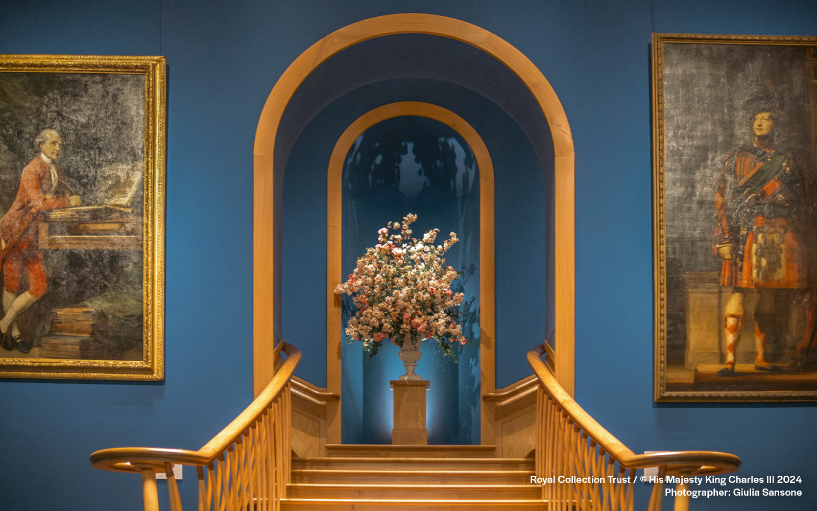 Floral arrangement on pedestal between two historical paintings in a blue-walled gallery.