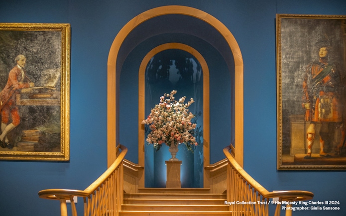 Floral arrangement on pedestal between two historical paintings in a blue-walled gallery.