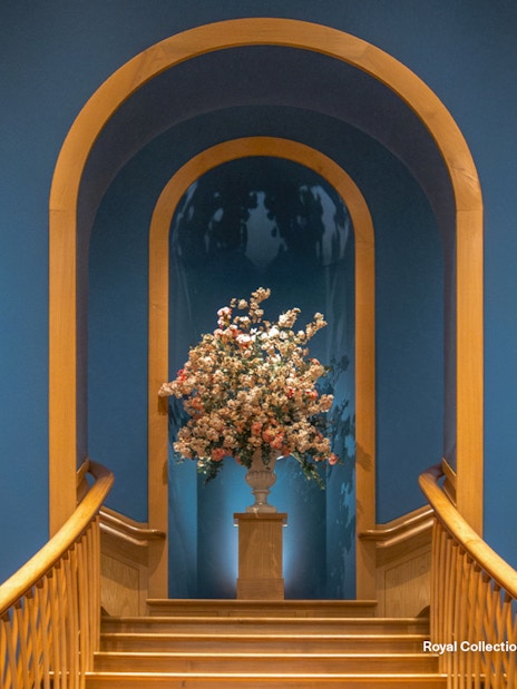 Floral arrangement on pedestal between two historical paintings in a blue-walled gallery.