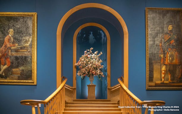 Floral arrangement on pedestal between two historical paintings in a blue-walled gallery.