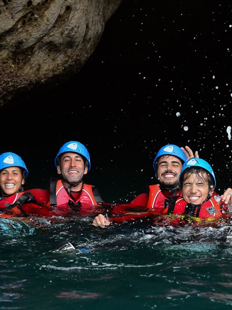 Group enjoying coasteering adventure in helmets and life jackets, splashing in water.