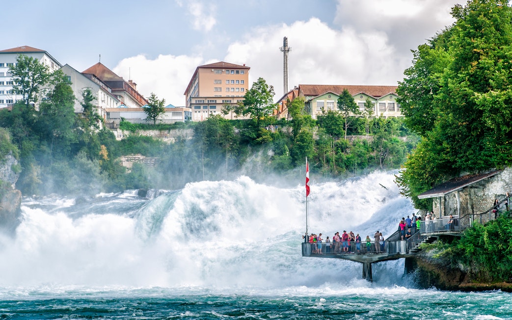 People on viewing platform at Rhine Falls, Switzerland, with waterfall and buildings in background.