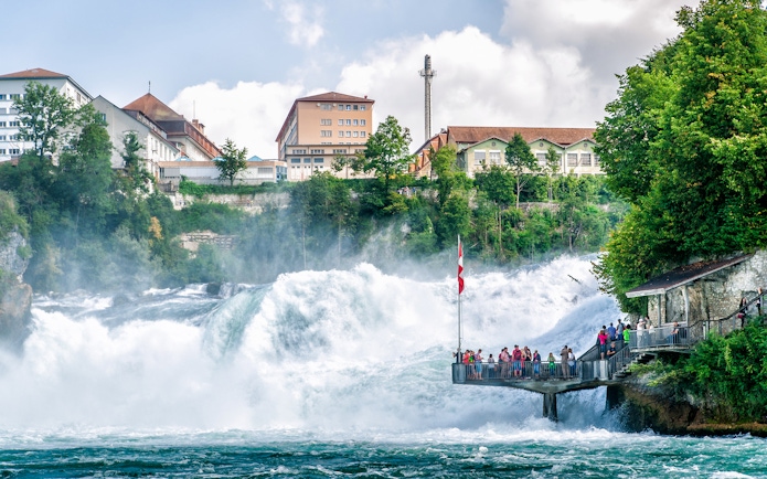 People on viewing platform at Rhine Falls, Switzerland, with waterfall and buildings in background.