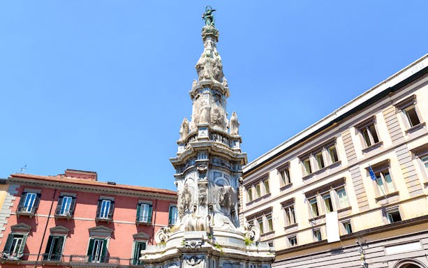 Baroque obelisk in Naples city square, Italy, with historic buildings in the background.