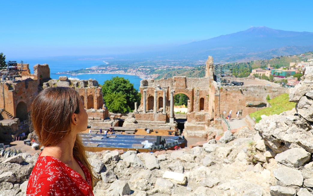 Young woman at ancient Greek theater ruins in Taormina with Mount Etna and Ionian Sea in the background.