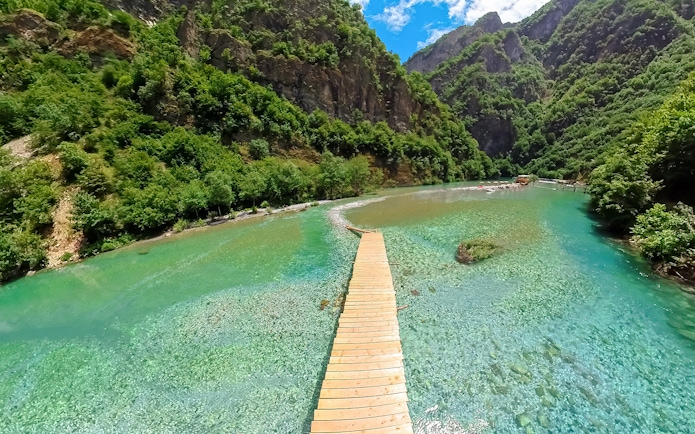 Wooden walkway over Shala River with lush green mountains during Komani Lake tour from Tirana.