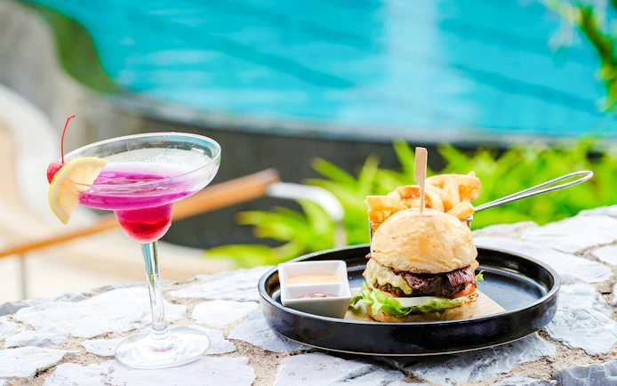 Burger and fries with a cocktail by a poolside in a tropical setting.