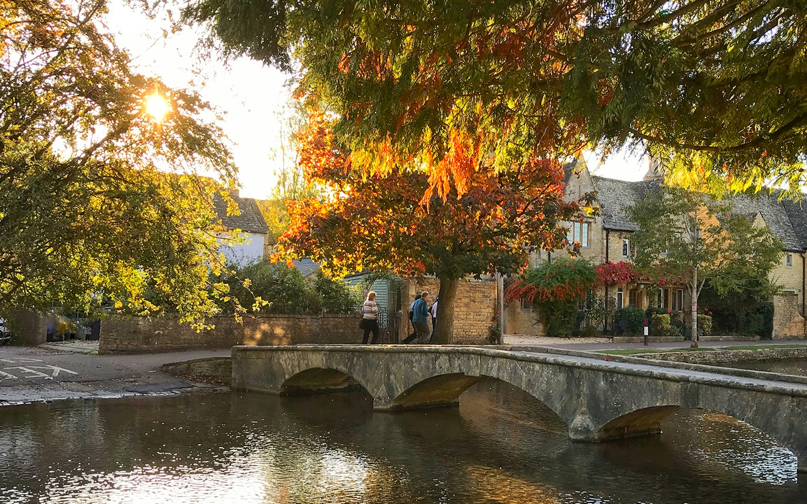 Stone bridge over river in Bourton-on-the-Water, Cotswolds, with autumn trees and historic buildings.