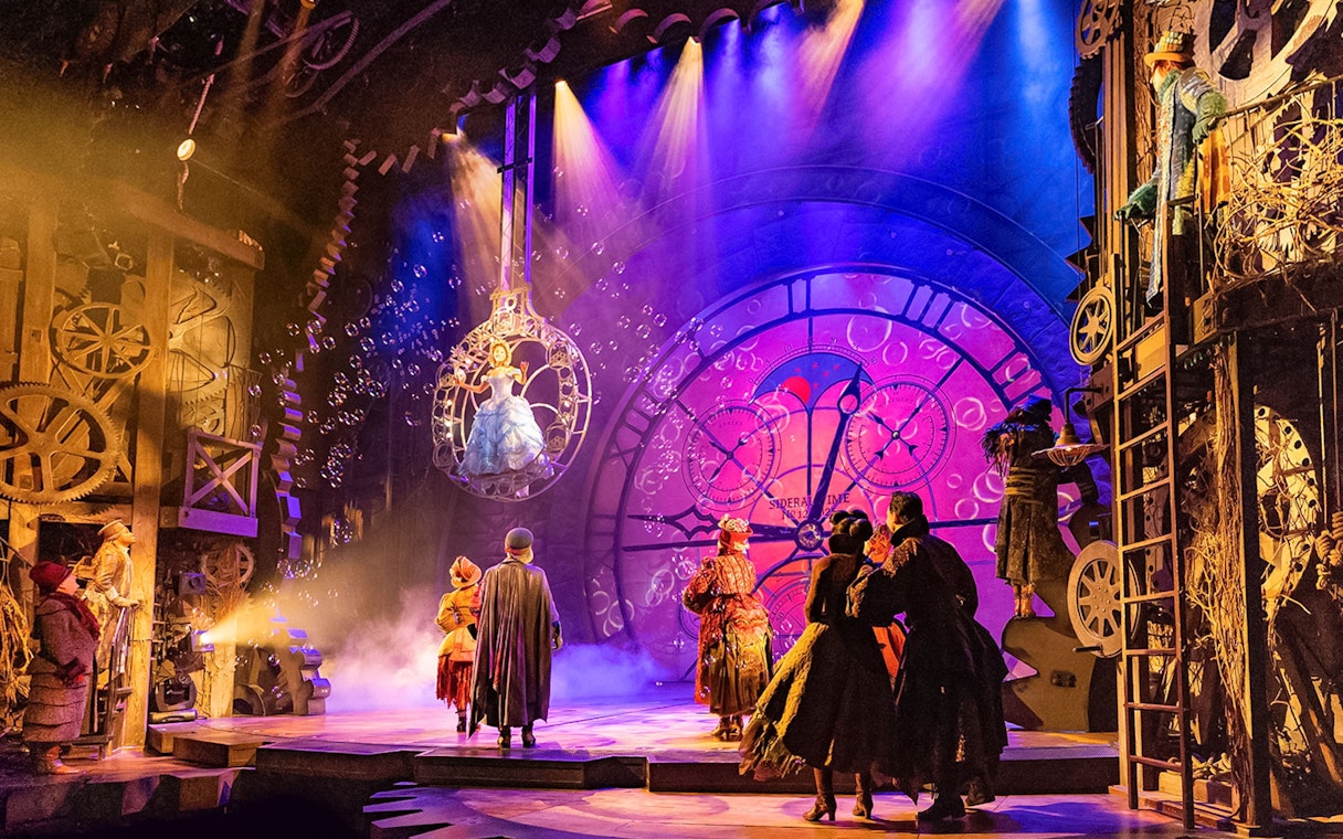Stage scene from the musical Wicked with performers in elaborate costumes and a large clock backdrop.