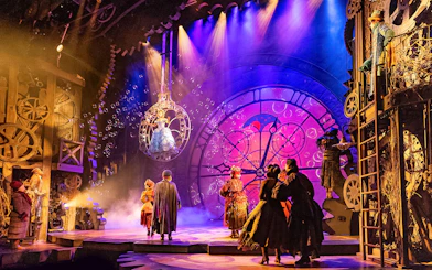 Stage scene from the musical Wicked with performers in elaborate costumes and a large clock backdrop.