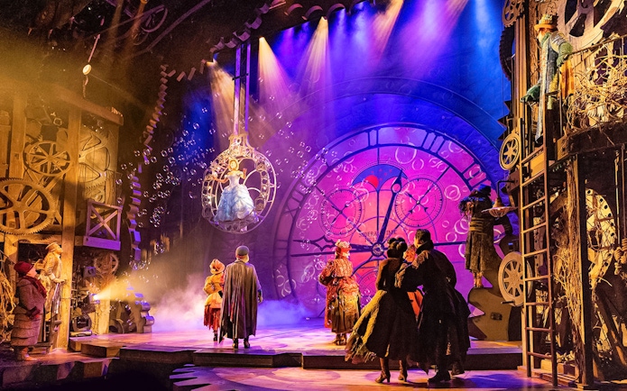 Stage scene from the musical Wicked with performers in elaborate costumes and a large clock backdrop.