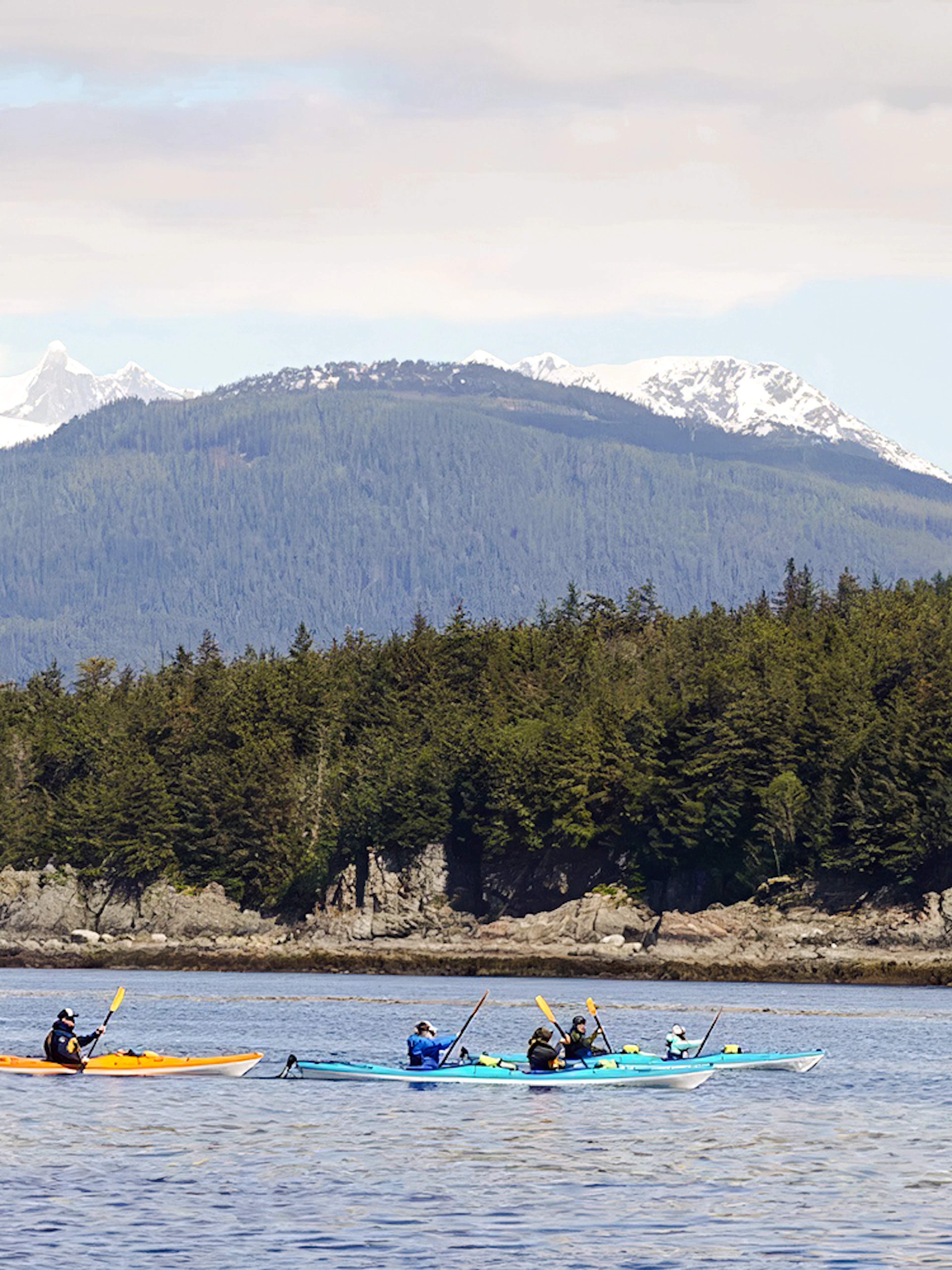 Kayakers paddling near forested coastline with snowy mountains in Juneau.