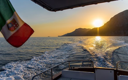 Boat cruising along the Amalfi Coast at sunset with Italian flag.