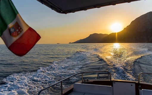 Boat cruising along the Amalfi Coast at sunset with Italian flag.