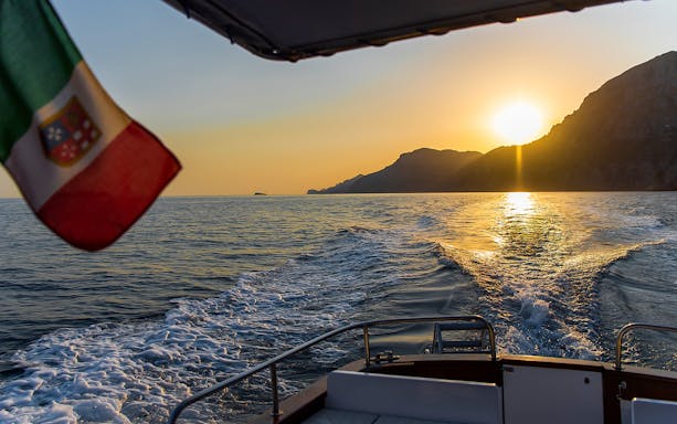 Boat cruising along the Amalfi Coast at sunset with Italian flag.