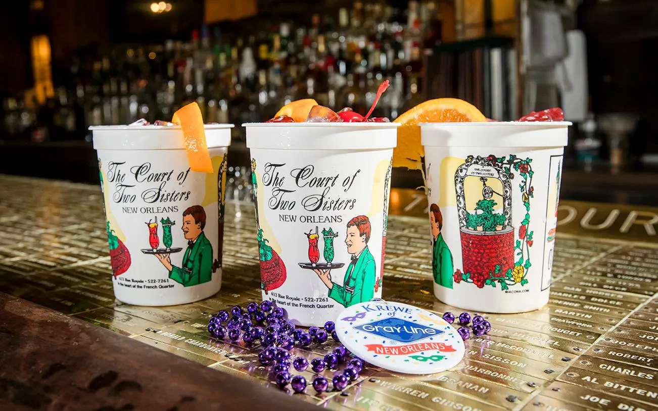 Three craft cocktails in branded cups at The Court of Two Sisters, New Orleans, with beads and a tour badge.