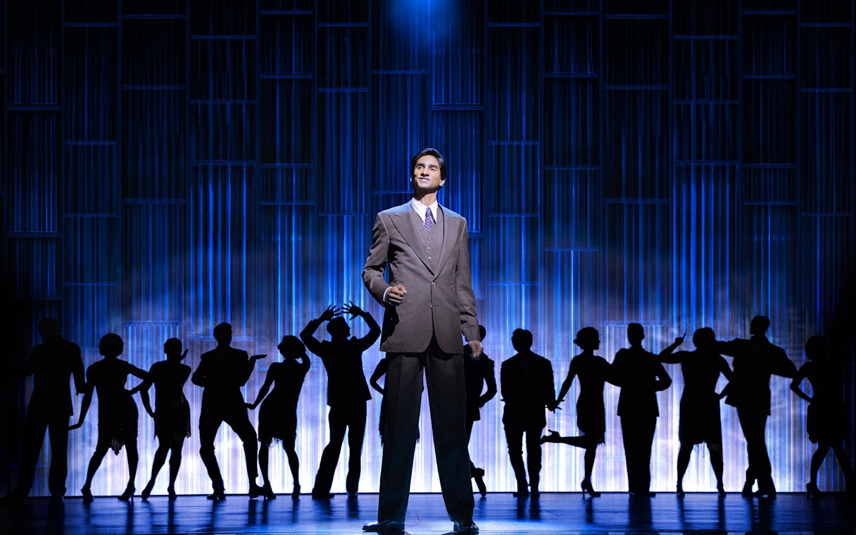 Man in suit on stage with dancers silhouetted against blue backdrop, The Great Gatsby performance.