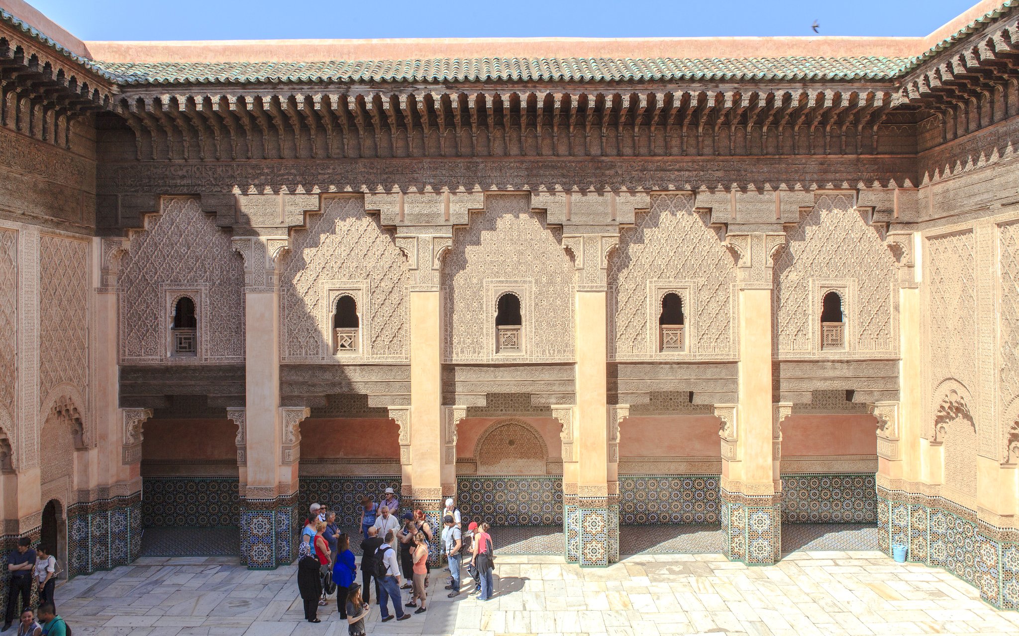 Courtyard of Ben Youssef Madrasa in Marrakech with intricate carvings and tourists exploring.