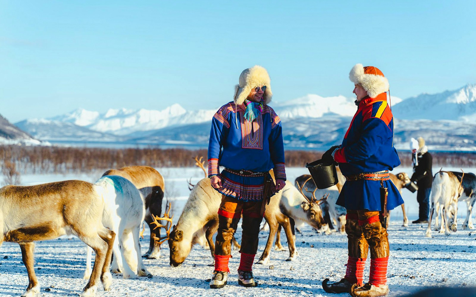 Sami people in traditional attire with reindeer in snowy landscape.
