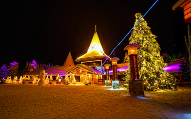 Santa Claus Village in Rovaniemi illuminated at night with snow-covered trees and festive lights.