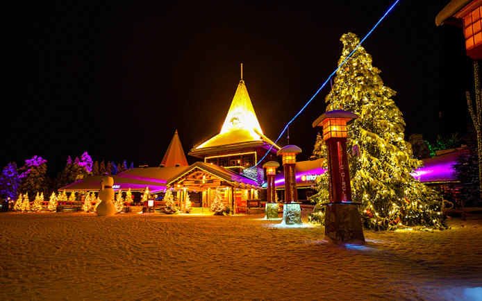 Santa Claus Village in Rovaniemi illuminated at night with snow-covered trees and festive lights.