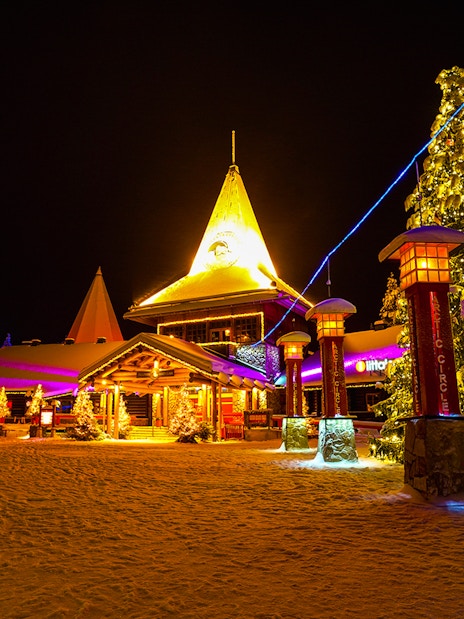 Santa Claus Village in Rovaniemi illuminated at night with snow-covered trees and festive lights.