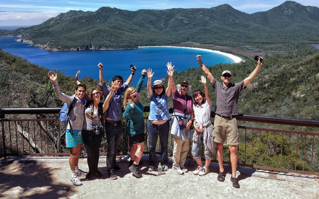 Tour group celebrating at Wineglass Bay lookout with ocean and mountains in background.