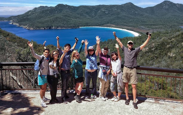 Tour group celebrating at Wineglass Bay lookout with ocean and mountains in background.