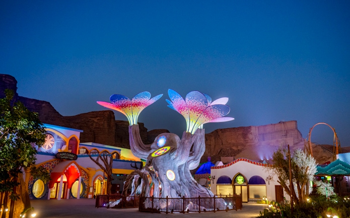 Colorful illuminated tree sculpture at Six Flags Qiddiya City against a twilight sky.