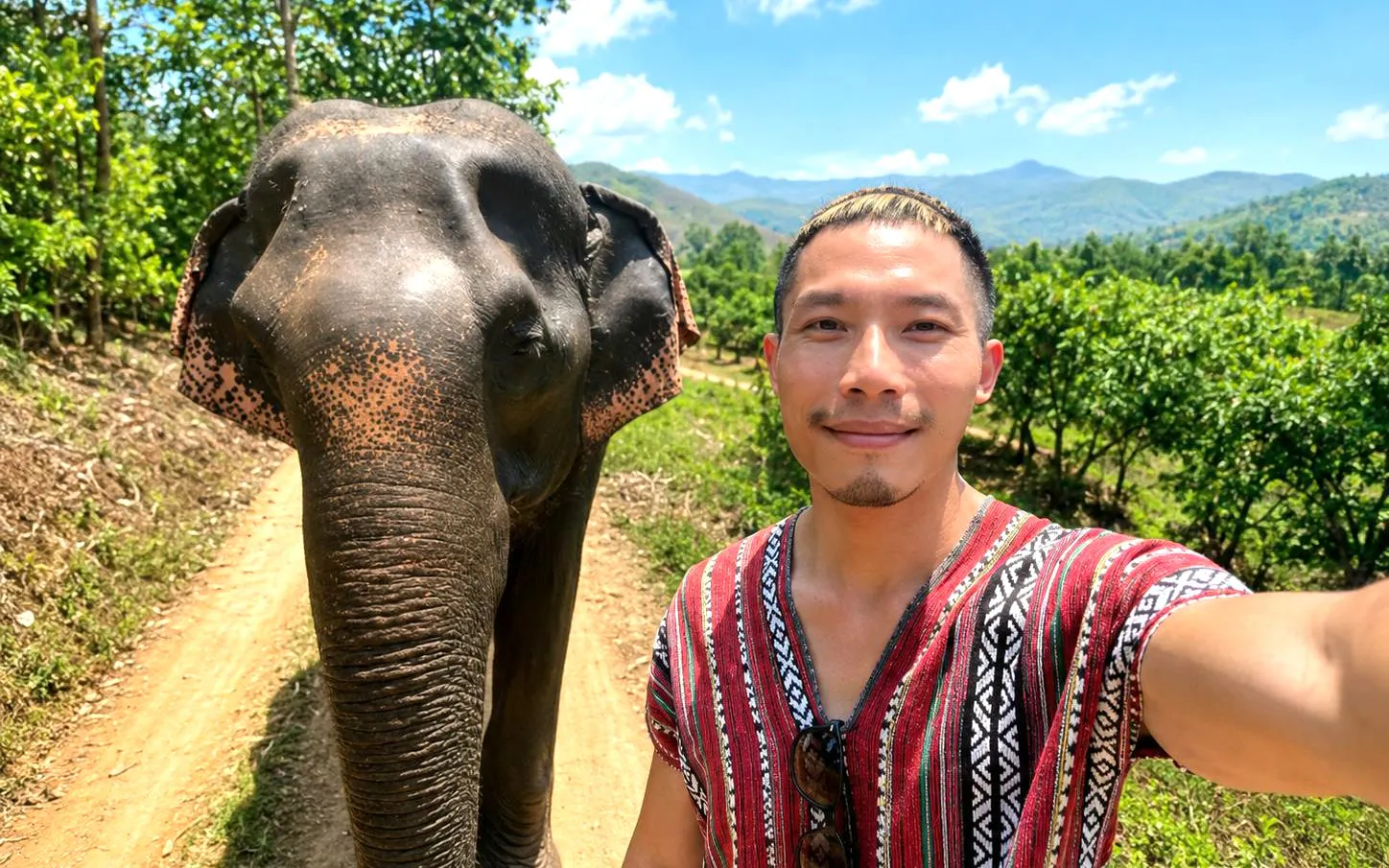 Man in traditional tunic with elephant on dirt path, Chiang Mai Elephant Care, Thailand.