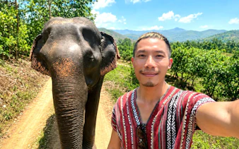 Man in traditional tunic with elephant on dirt path, Chiang Mai Elephant Care, Thailand.