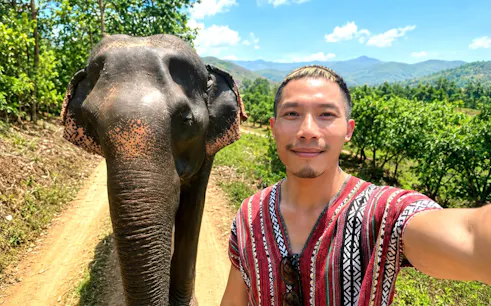Man in traditional tunic with elephant on dirt path, Chiang Mai Elephant Care, Thailand.