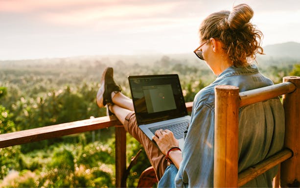Woman using a laptop on a balcony overlooking a lush landscape.