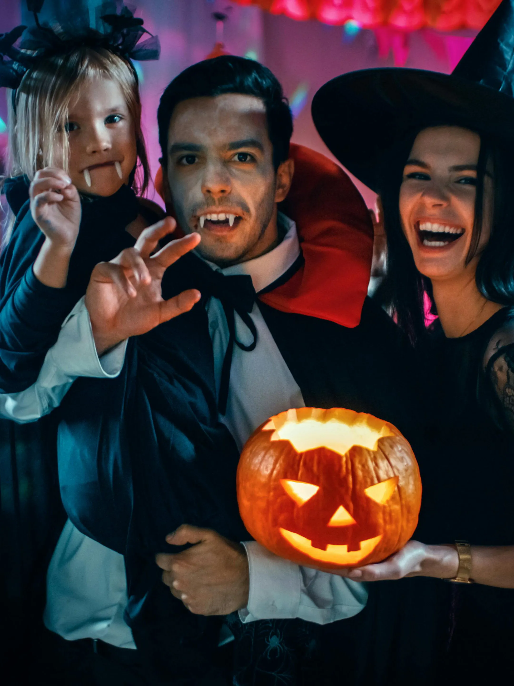 Family in vampire and witch costumes holding a carved pumpkin at a Halloween party.