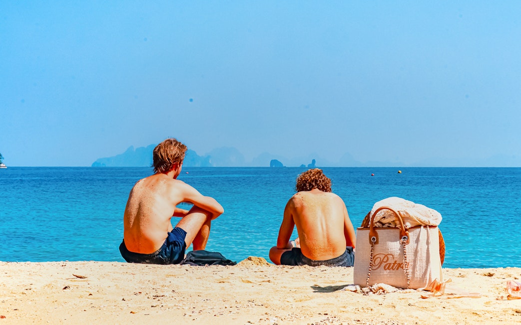 Two people sitting on a sandy beach in Krabi, Thailand, facing the ocean.