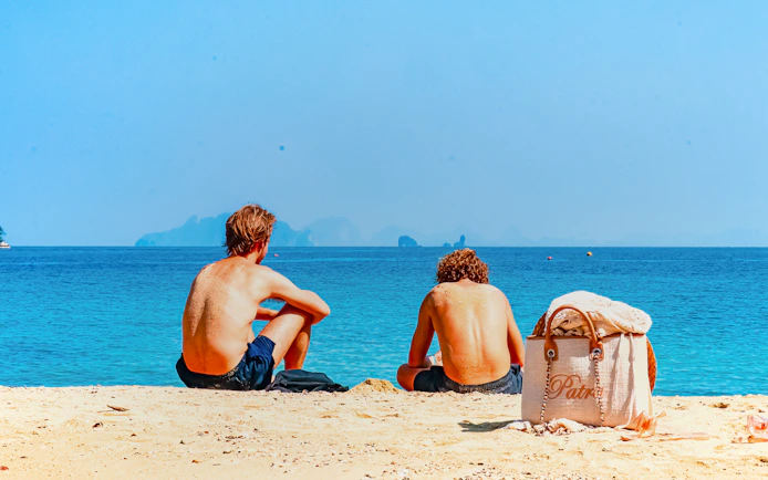 Two people sitting on a sandy beach in Krabi, Thailand, facing the ocean.