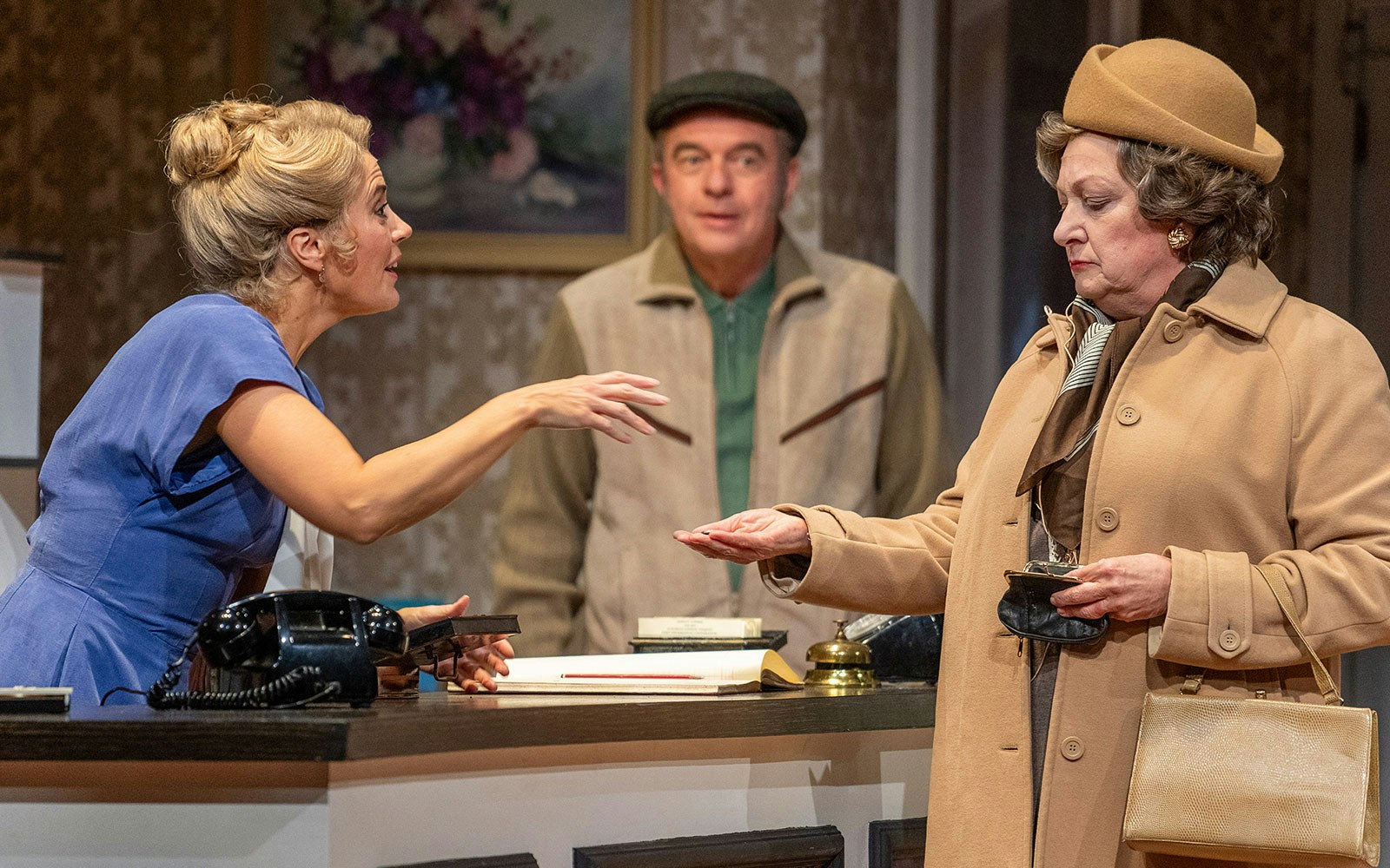 Receptionist interacting with guests at Fawlty Towers hotel desk.