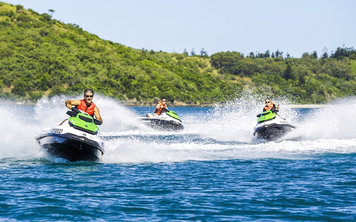 Tourists riding jetskis in the Whitsundays with lush green hills in the background.