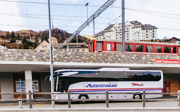 Bernina Red Train passing through St. Moritz station with a tour bus parked below.