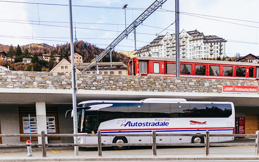 Bernina Red Train passing through St. Moritz station with a tour bus parked below.